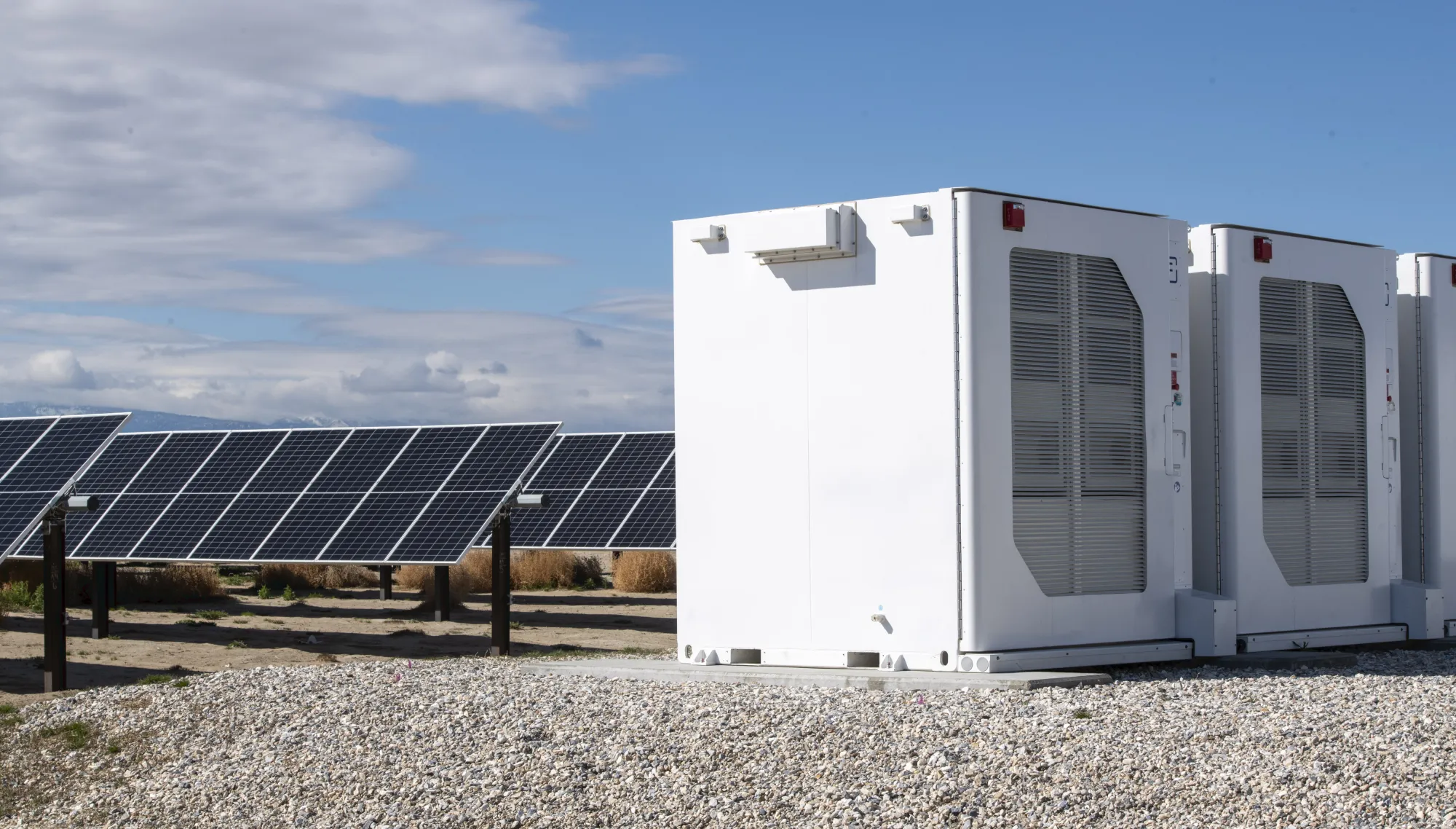 A row of solar panels is positioned on the left side of the image, set against a clear blue sky with some clouds. On the right side, there are large white industrial units with vents, situated on a gravel surface.