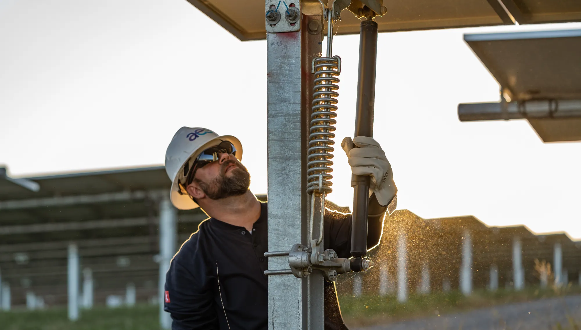 A worker wearing a hard hat and safety glasses adjusts a solar panel mechanism at a solar farm. The sun is setting in the background, casting a warm glow on the scene. The worker is holding a tool and wearing gloves, indicating a focus on safety.