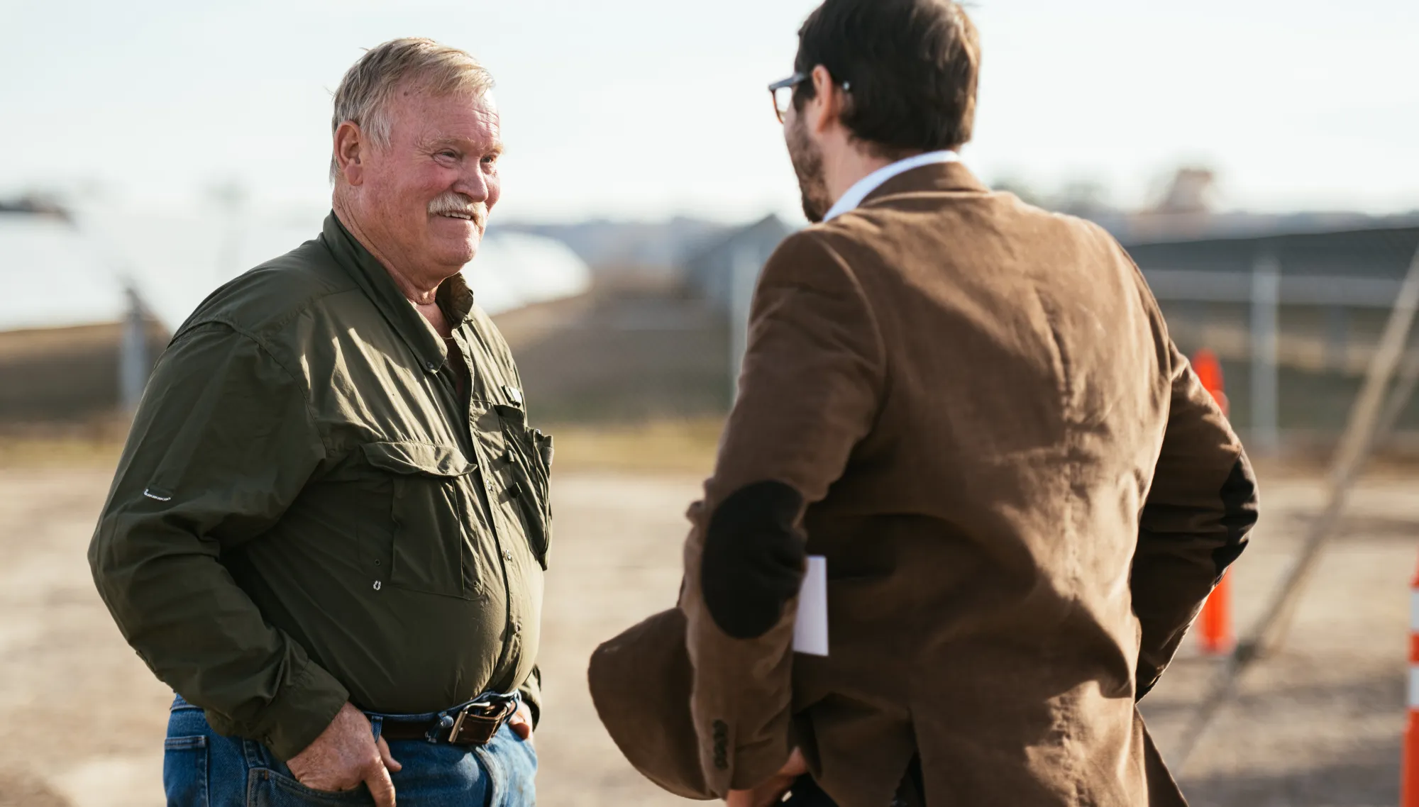 Two men having a conversation outdoors, one wearing a green shirt and jeans, the other in a brown jacket with elbow patches. They are standing in front of a blurred background with fencing and construction cones.