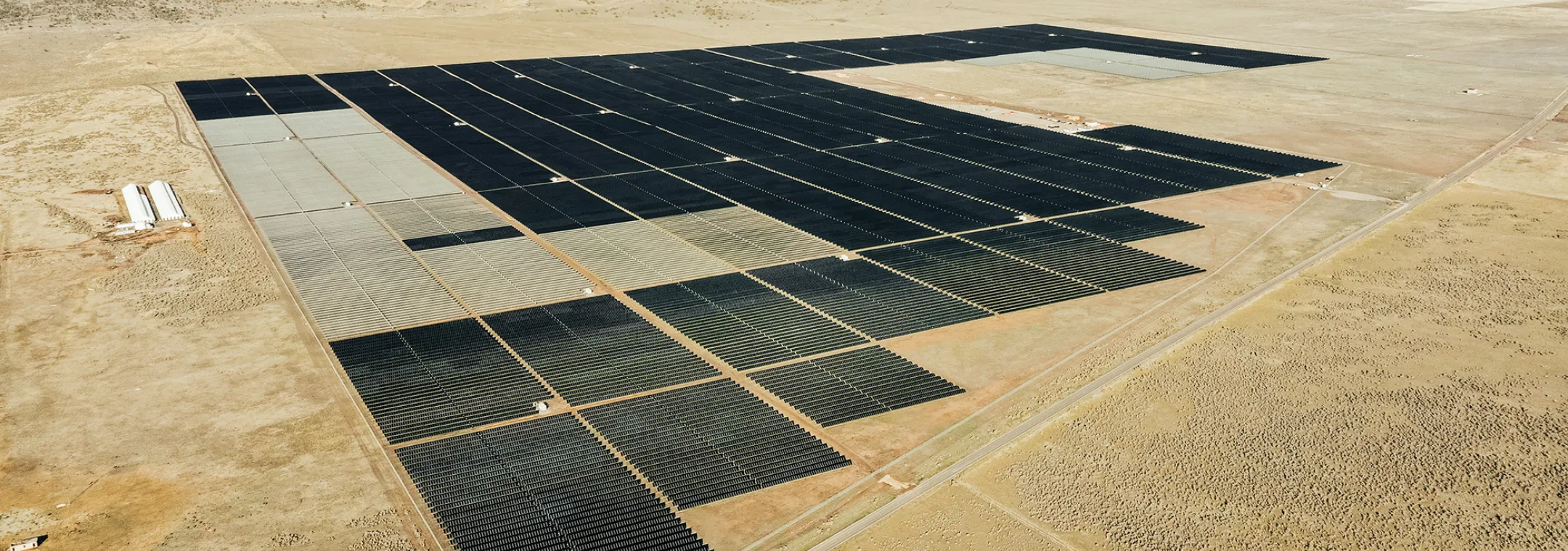 Aerial view of a large solar farm in a desert landscape, with rows of solar panels stretching across the dry, flat terrain. Mountains are visible in the background under a clear sky.