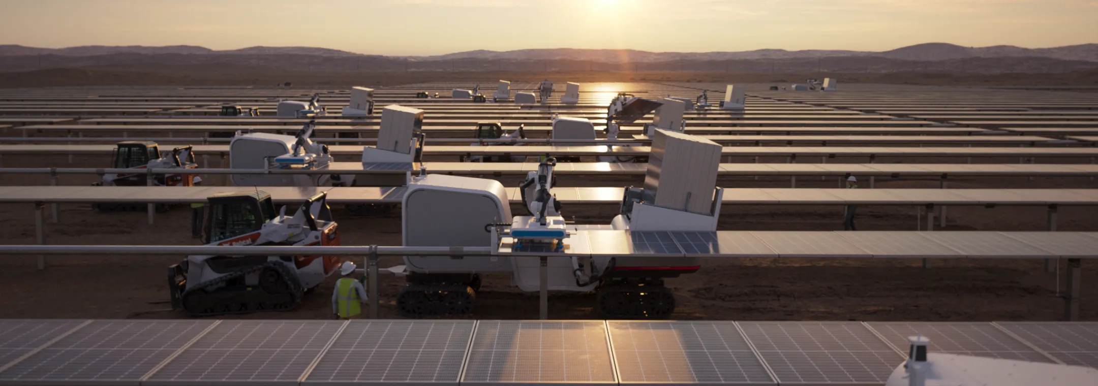 A large solar farm at sunset with rows of solar panels and machinery. The sun is low on the horizon, casting a warm glow over the landscape. Several pieces of equipment, including a Bobcat, are visible among the panels.