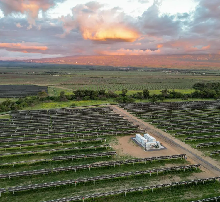 Aerial view of a solar farm with rows of solar panels under a cloudy sky at sunset. The landscape is green with some trees, and there are mountains in the background.