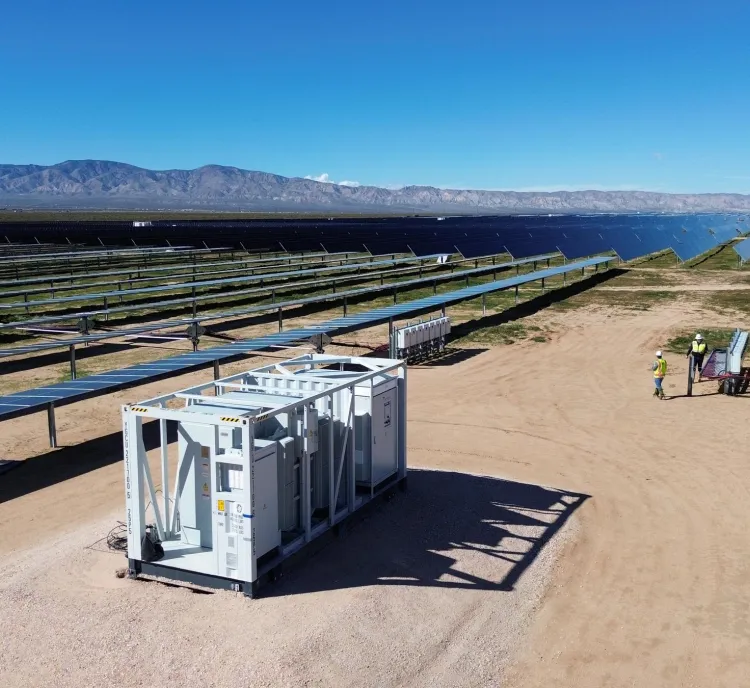Aerial view of a large solar farm with rows of solar panels extending into the distance under a clear blue sky. A white utility structure is in the foreground, and two workers in safety vests are inspecting equipment nearby.