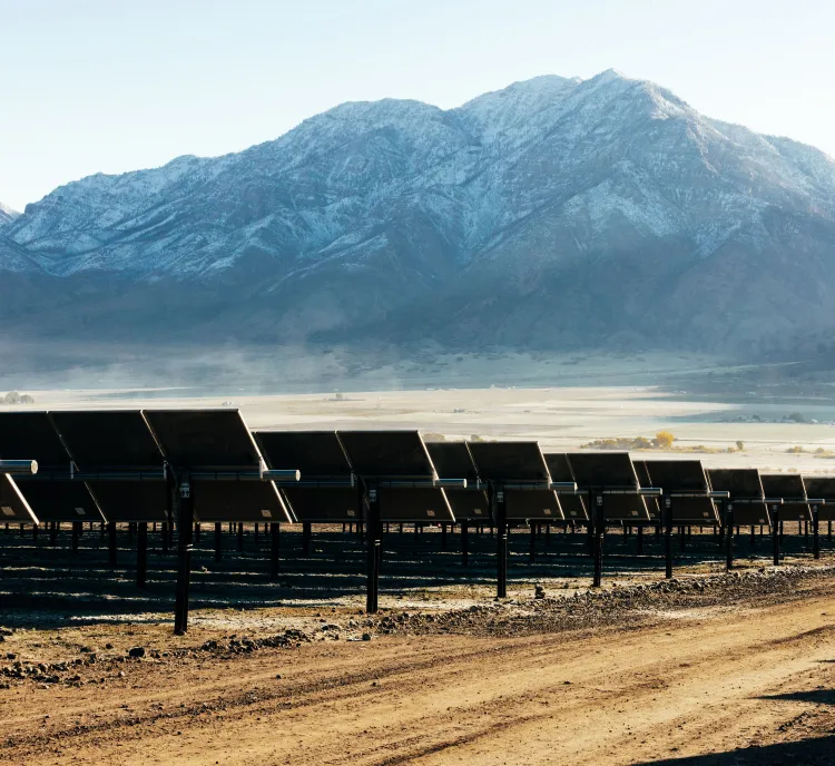 Rows of solar panels on a dirt field with snow-capped mountains in the background under a clear blue sky.