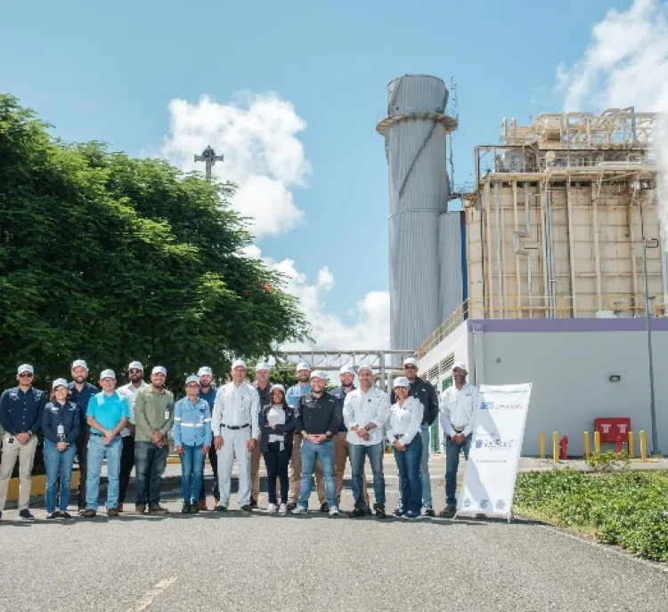 A group of people wearing hard hats and safety gear stand in front of an industrial facility with large structures emitting steam. The sky is clear with some clouds, and there is a tree on the left side of the image. A signboard with text is visible on the right.