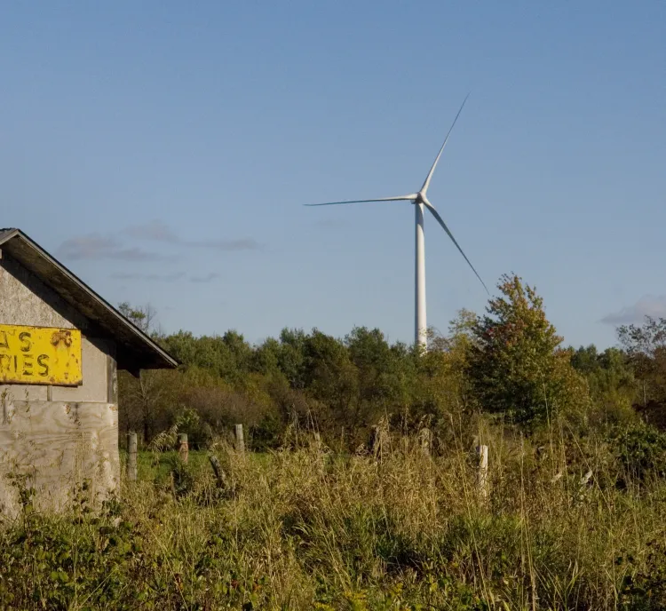 A rustic wooden building with a yellow sign reading 'Bova's Groceries' stands in a grassy field. In the background, a wind turbine rises above a line of trees against a clear blue sky.
