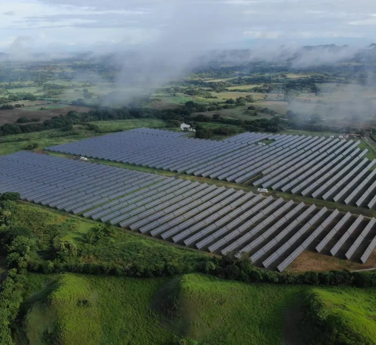 Aerial view of a large solar farm with rows of solar panels surrounded by lush green fields and hills. Misty clouds hover above the landscape, and distant mountains are visible under a cloudy sky.