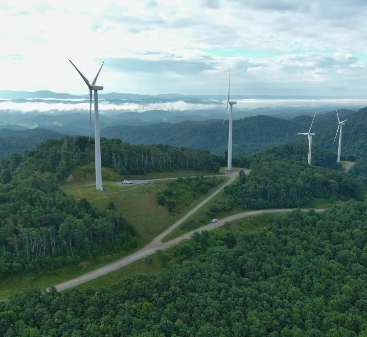 Aerial view of a lush green landscape with several wind turbines on hilltops. A winding road passes through the hills, and clouds hover over distant mountains under a partly cloudy sky.