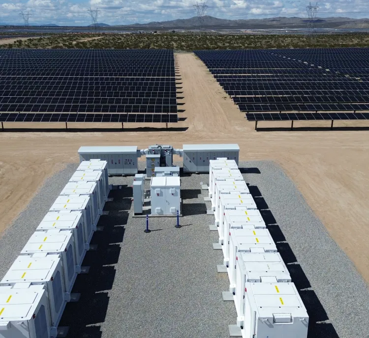Aerial view of a solar farm with rows of solar panels and white utility boxes on a gravel path, set in a desert landscape under a cloudy sky.