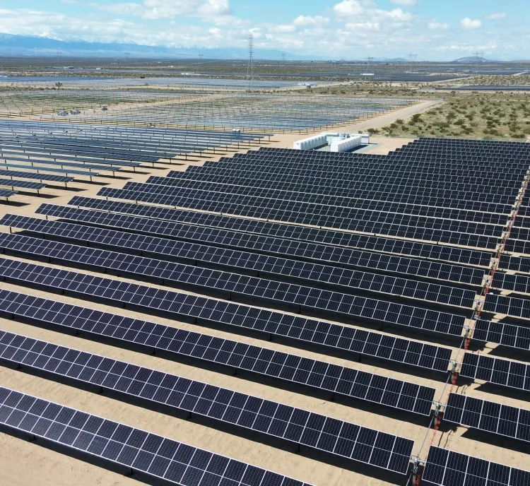 Aerial view of a large solar farm with rows of solar panels stretching across a desert landscape. Mountains are visible in the distant background under a partly cloudy sky.
