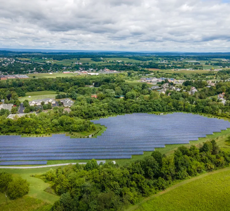 Aerial view of a large solar panel farm surrounded by green fields and trees. In the background, there are residential areas with houses and roads, set against a backdrop of rolling hills and a cloudy sky.