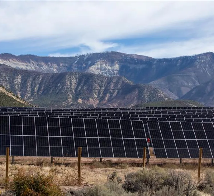 A field of solar panels is set against a backdrop of rugged mountains under a partly cloudy sky. The landscape has sparse vegetation and a fence surrounding the solar array.