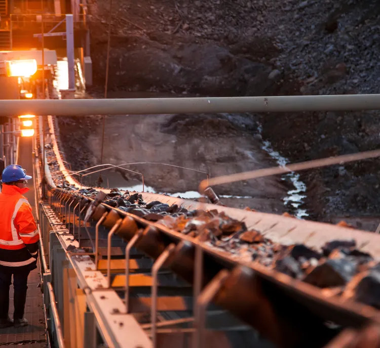 A worker in a bright orange safety jacket and blue helmet stands next to a conveyor belt carrying rocks at an industrial mining site during sunset.