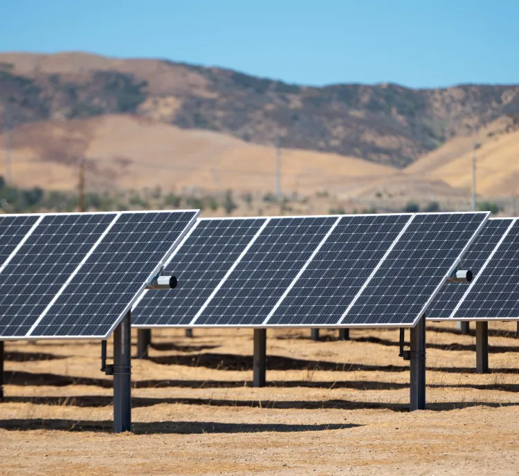 Rows of solar panels in a dry landscape with hills in the background under a clear blue sky.