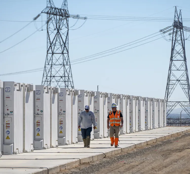 Two workers walk along a row of large white battery storage units with power lines and towers in the background under a clear blue sky.
