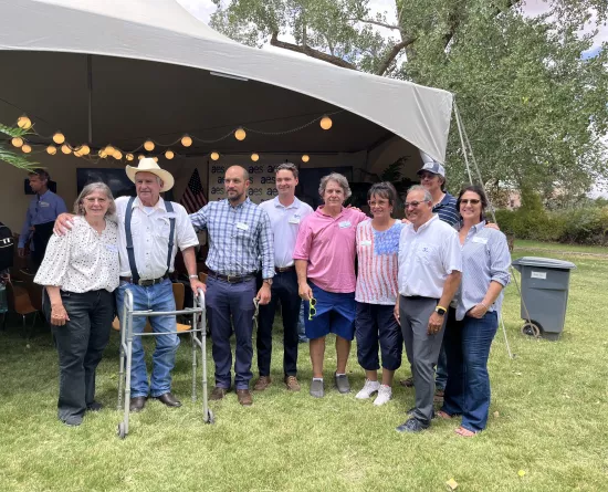 A group of nine people standing on grass in front of a tent. Some are wearing casual clothes like jeans and shirts, while others are dressed in business casual attire. One person uses a walker. The tent behind them is decorated with string lights and a banner with 'aes' text. Trees and a trash bin are visible in the background.