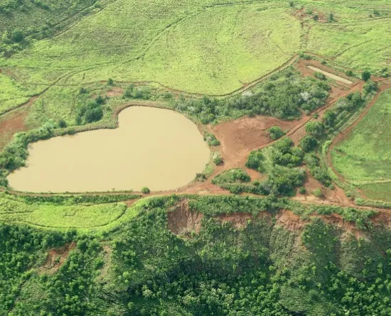 Aerial view of a green landscape with a heart-shaped pond in the center. The surrounding area has patches of grass, trees, and dirt paths. The landscape is divided into sections by natural and man-made boundaries.