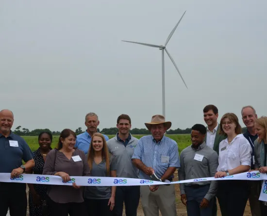 A group of people standing in front of a wind turbine, holding a ribbon labeled 'aes' during a ribbon-cutting ceremony in a field.