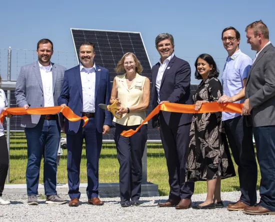 A group of people stand in front of solar panels, holding an orange ribbon at a ribbon-cutting ceremony. One person holds large scissors, ready to cut the ribbon. The event takes place outdoors under a clear blue sky.