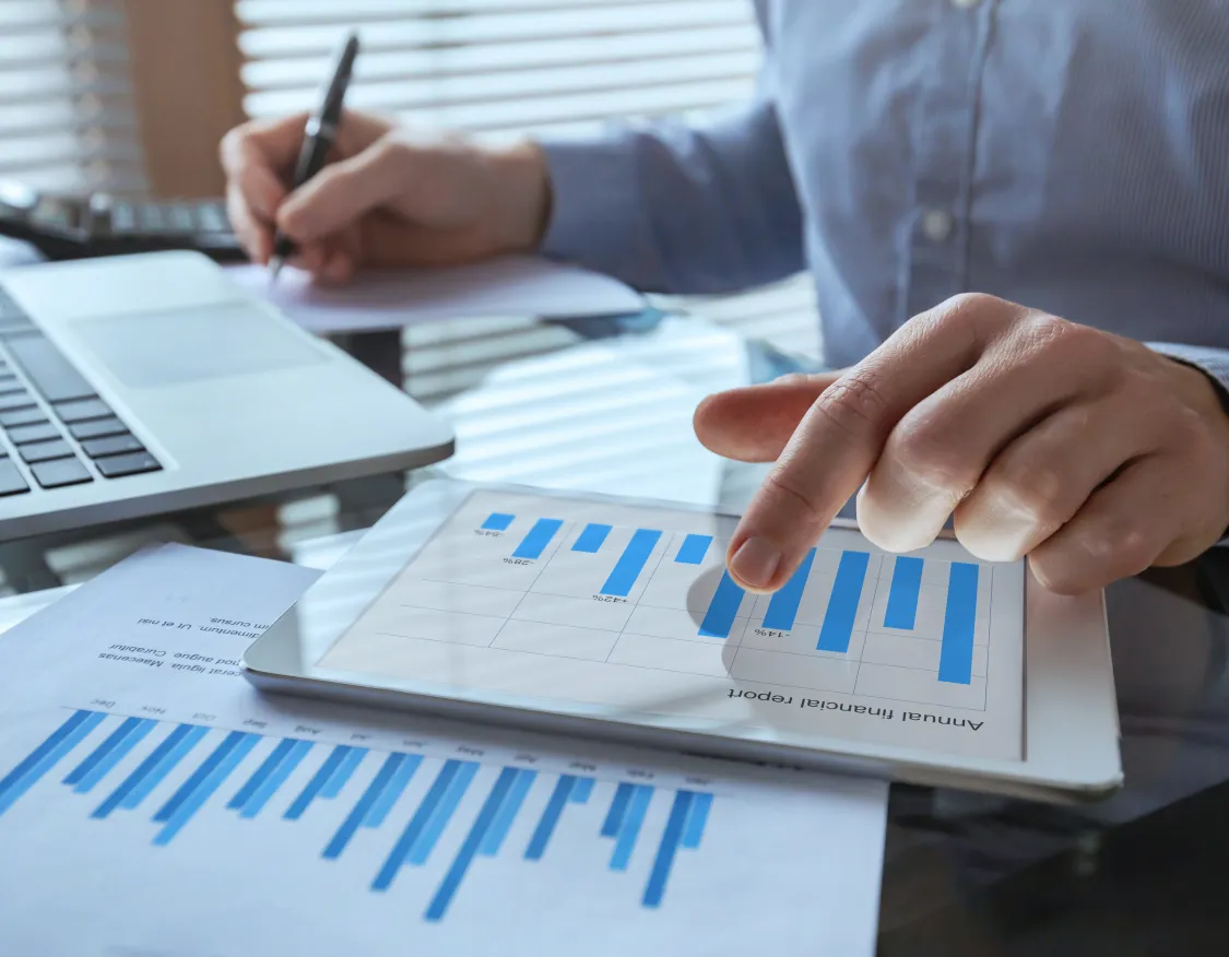 A person analyzing blue bar graphs on a tablet and paper, with a laptop and calculator nearby on a desk.