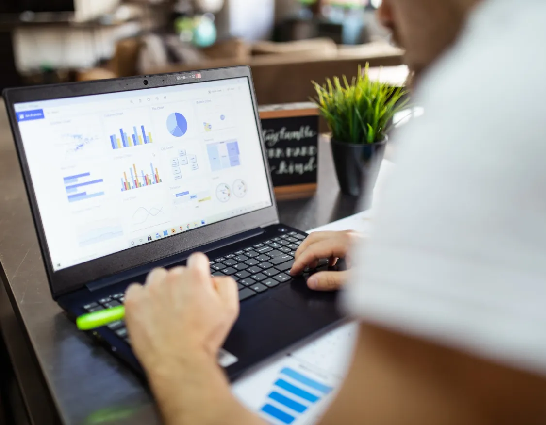 Person working on a laptop displaying various charts and graphs, with a potted plant and a sign in the background.