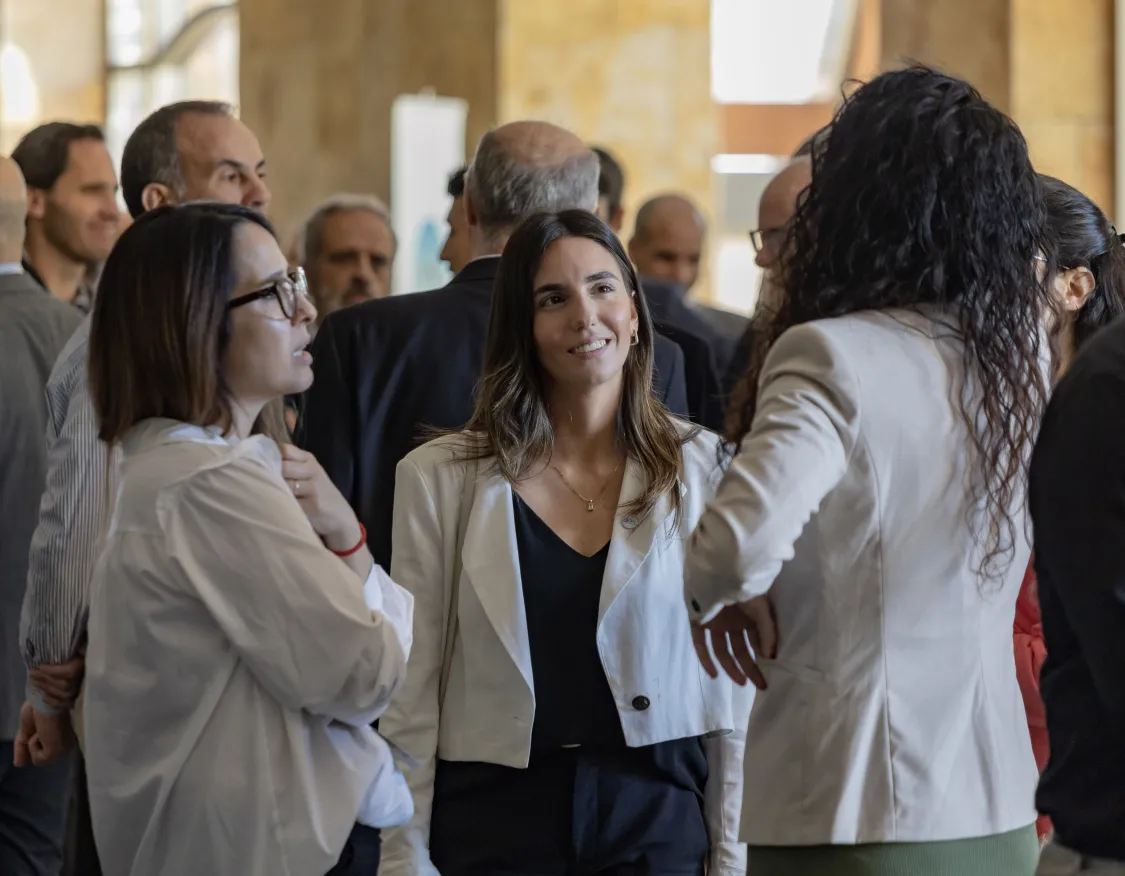 A group of people in business attire are engaged in conversation at an indoor event. The focus is on a woman in a white jacket smiling while talking to others. The background shows more people mingling in a well-lit room with large windows.