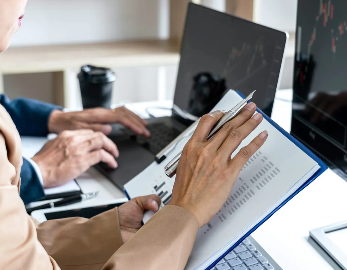 Two people working at a desk with laptops and financial charts on screens. One person is holding a pen and a document.