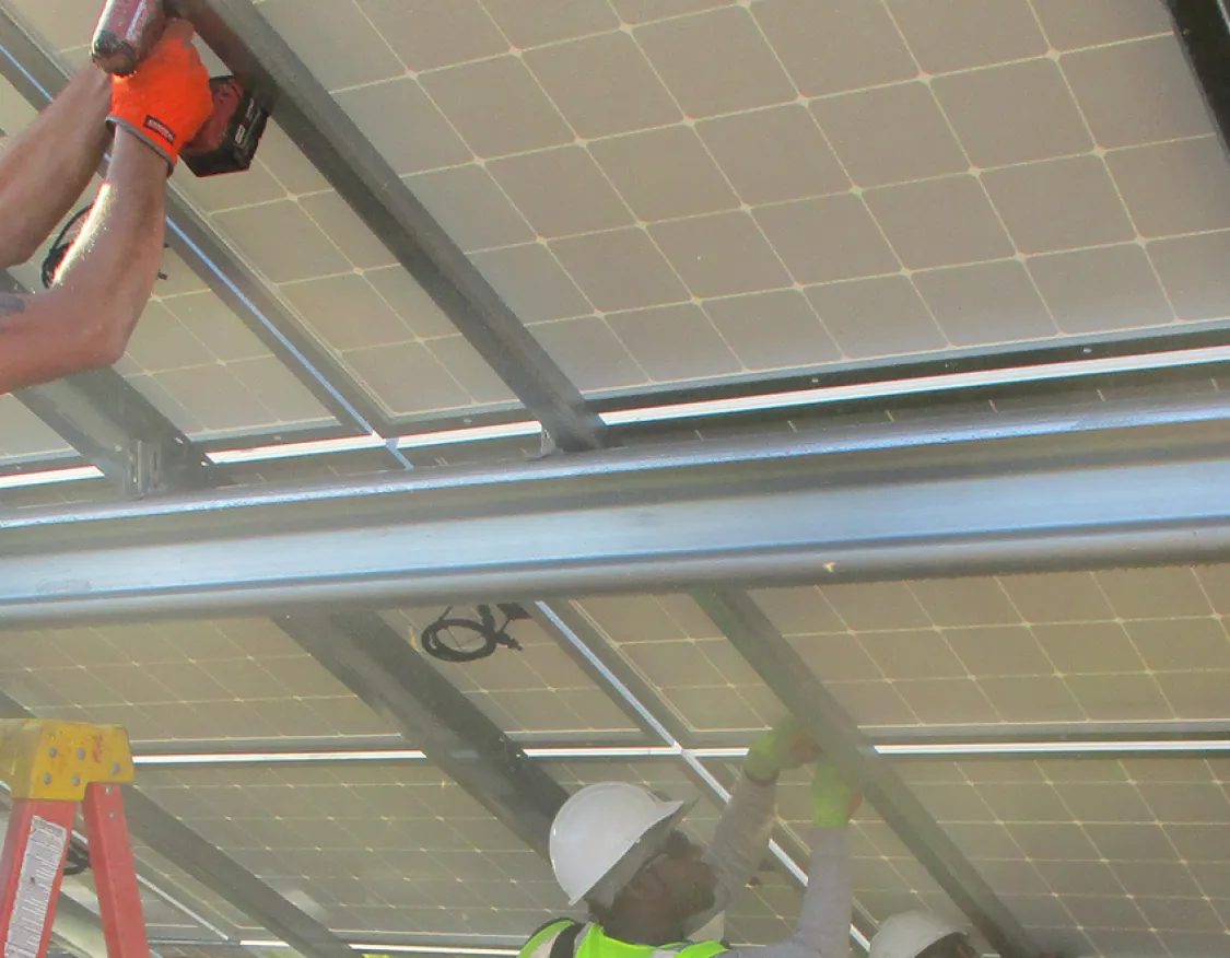 Construction workers wearing safety vests and helmets are installing solar panels on a metal structure. One worker is using a power drill, while others assist from below.