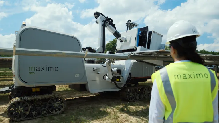 A person in a hard hat and high-visibility vest stands near a large robotic machine on tracks, labeled 'maximo aes', under a partly cloudy sky.