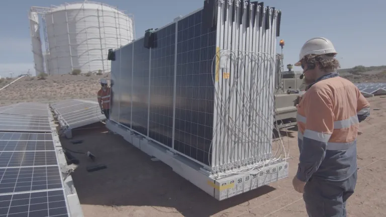 Two workers in orange safety gear and helmets stand near large solar panels in a desert area. A large white industrial tank is visible in the background.