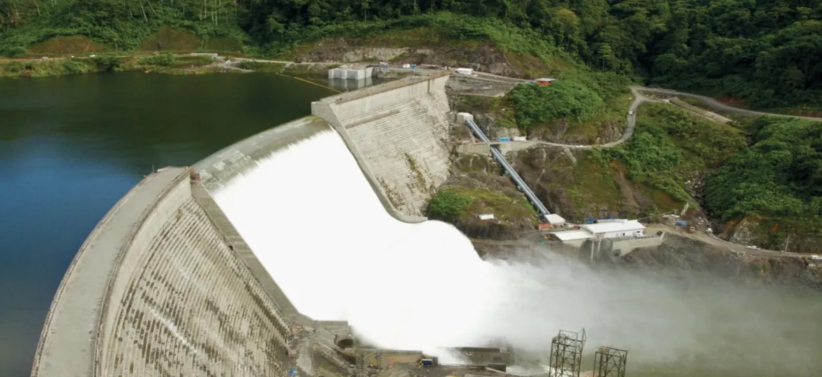 Aerial view of a large dam with water flowing over it, surrounded by lush green forest and a winding road.