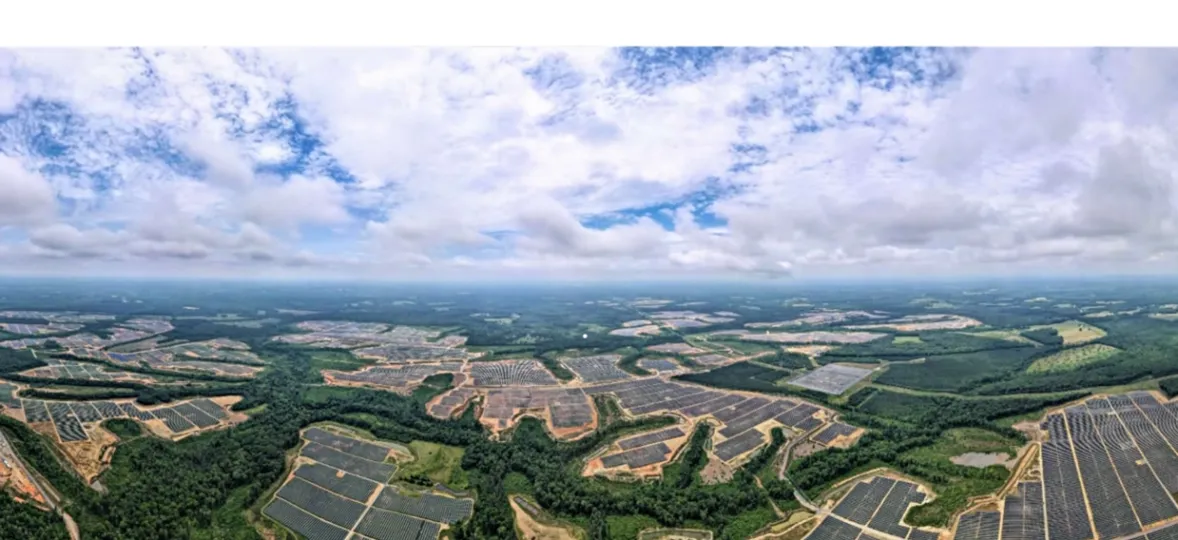 Aerial view of a large solar farm with numerous solar panels spread across a vast landscape, surrounded by green forests and under a partly cloudy sky.