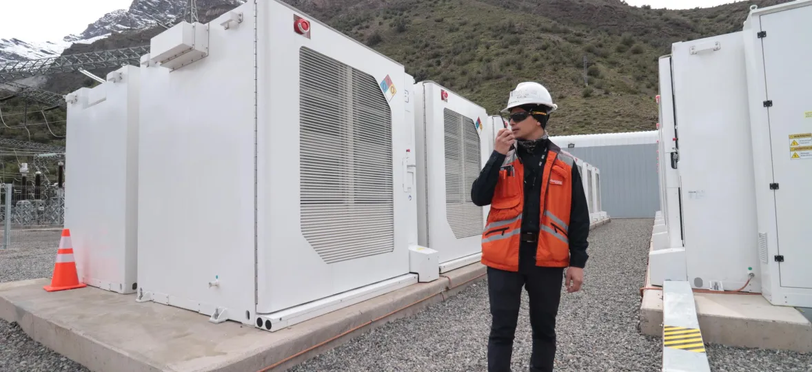 A worker in an orange safety vest and hard hat uses a walkie-talkie near large white industrial units. Snow-capped mountains are visible in the background.