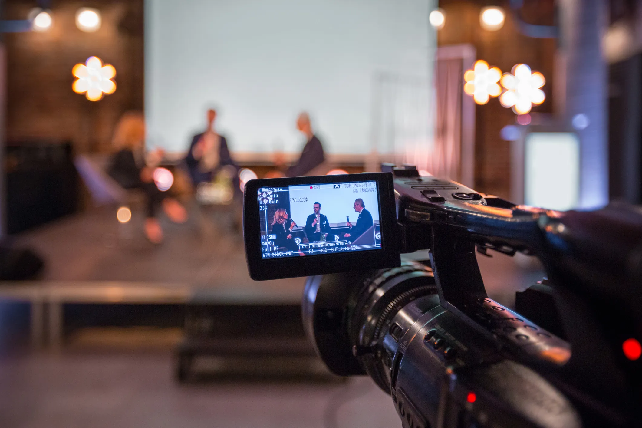 A video camera records a panel discussion on a stage, with two men and one woman seated and speaking. The camera's viewfinder shows a clear image of the panelists, while the background is blurred with warm lighting.