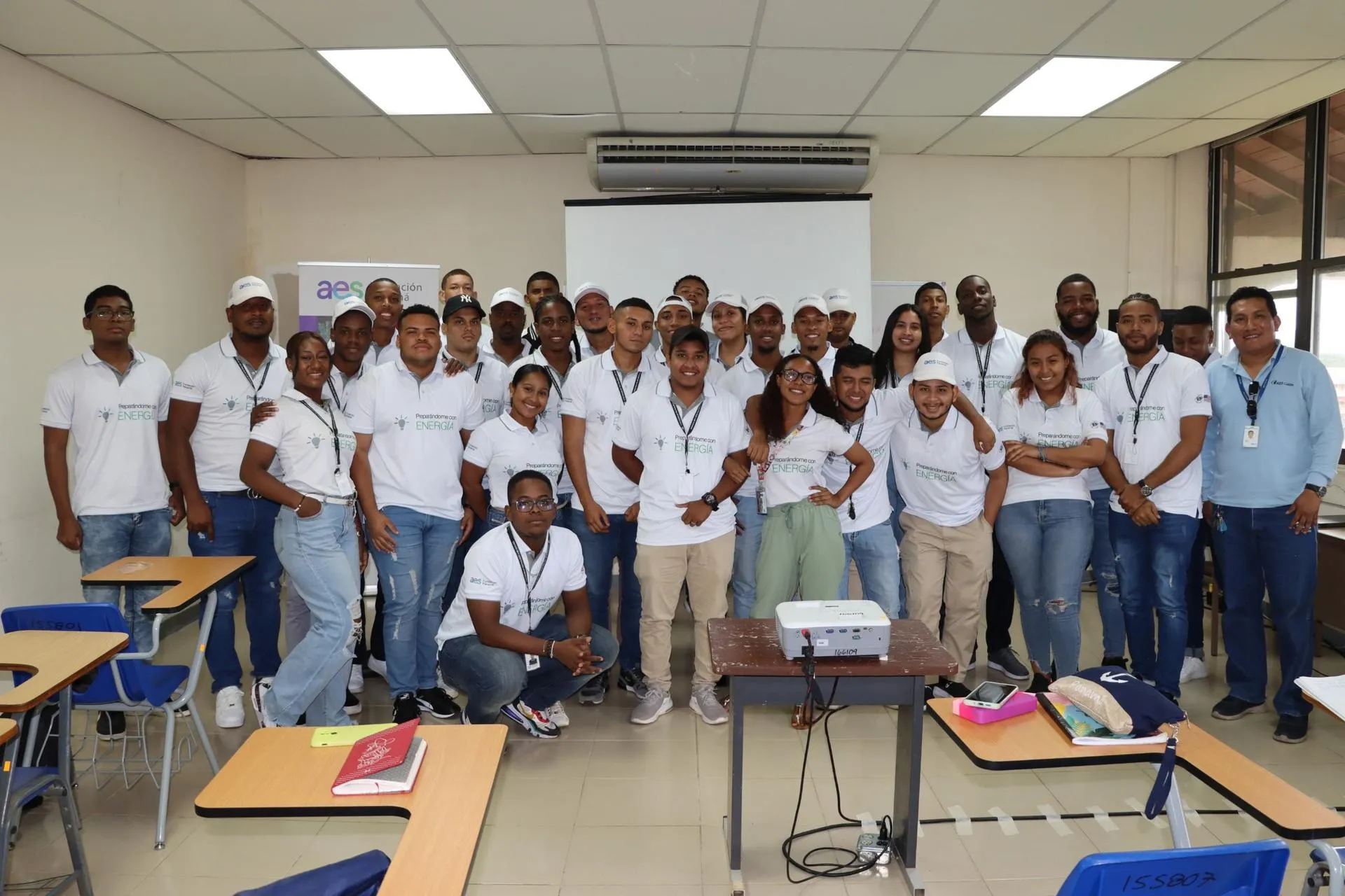A group of people in a classroom, wearing matching white shirts with a logo and text, pose for a group photo. They are standing and smiling, with a projector on a table in the foreground. Desks and chairs are visible around them.