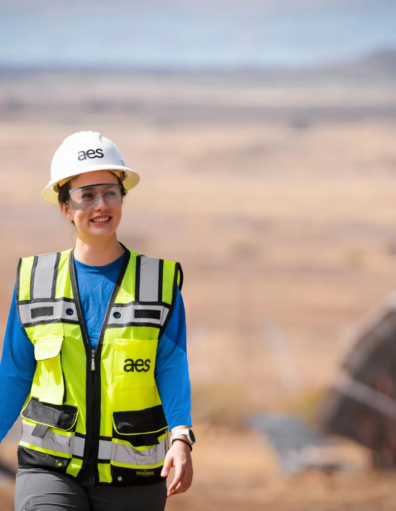 A person wearing a hard hat and safety vest smiles while walking through a solar panel field.