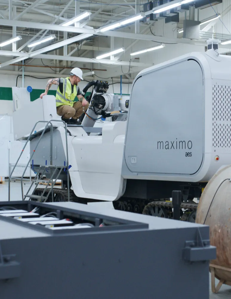 A worker in a safety vest and helmet operates a large industrial machine labeled 'maximo aes' inside a well-lit factory with exposed ceiling beams.
