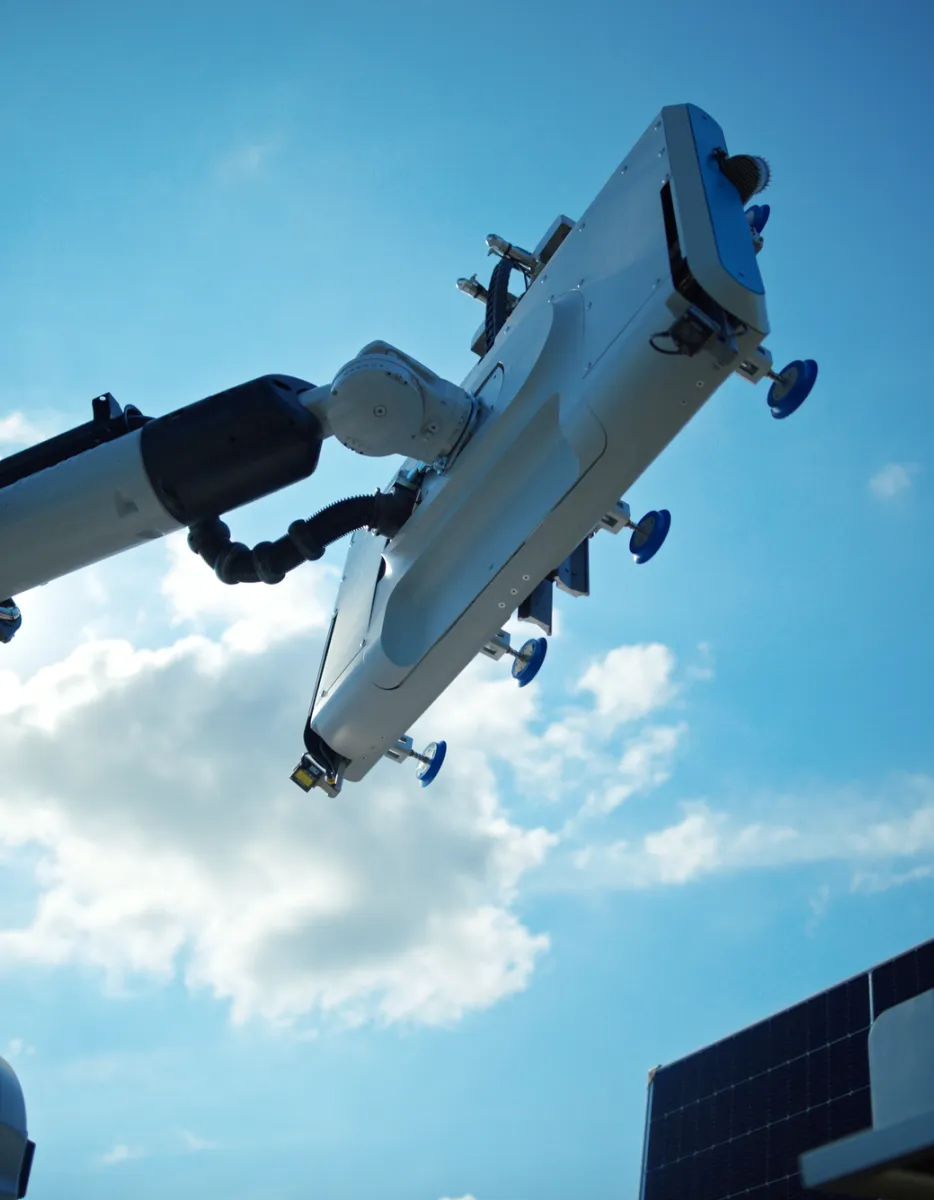 A robotic arm with suction cups is positioned against a bright blue sky with clouds, near solar panels.