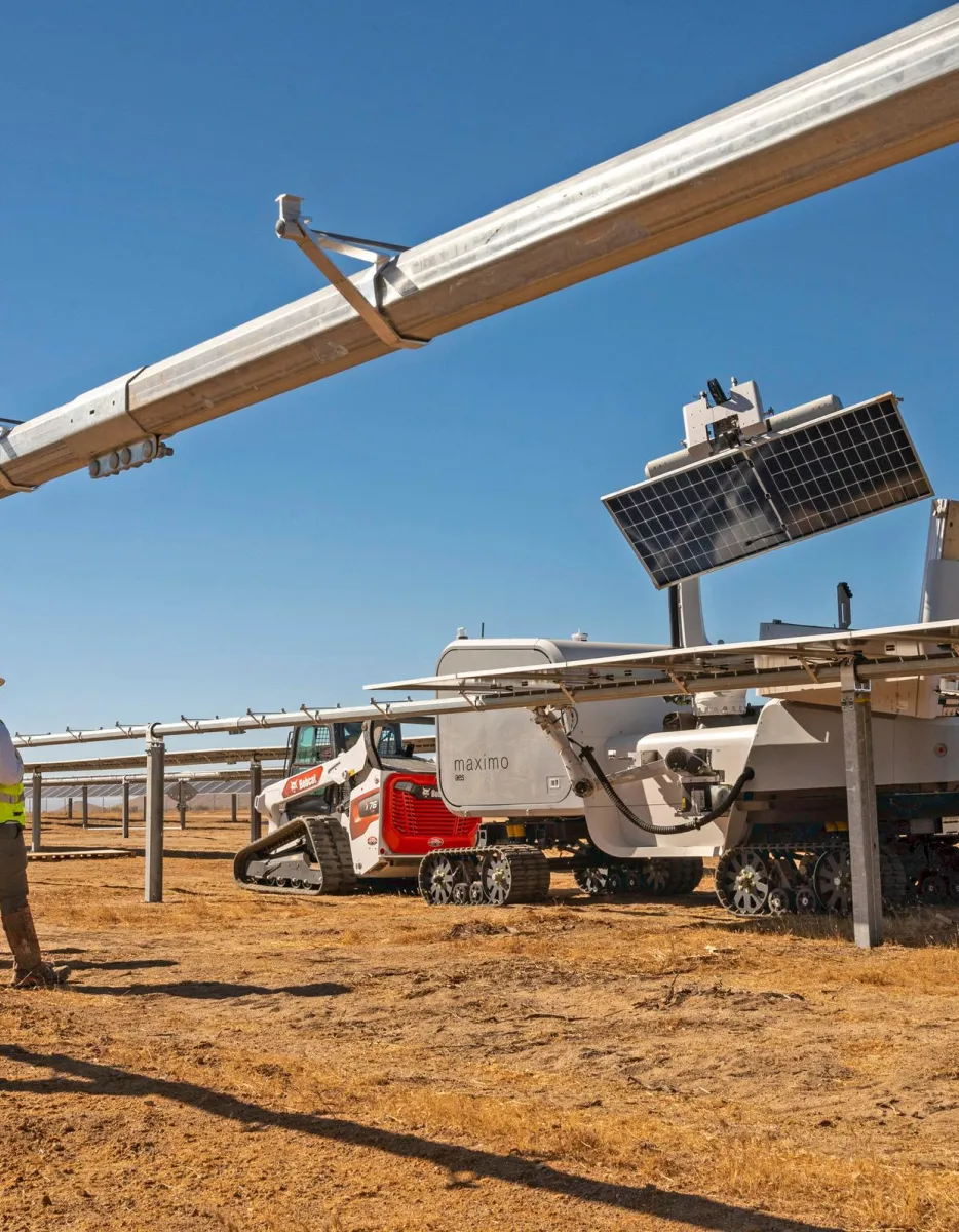 Three workers in safety gear stand near a solar panel installation site with a robotic vehicle on a sunny day.
