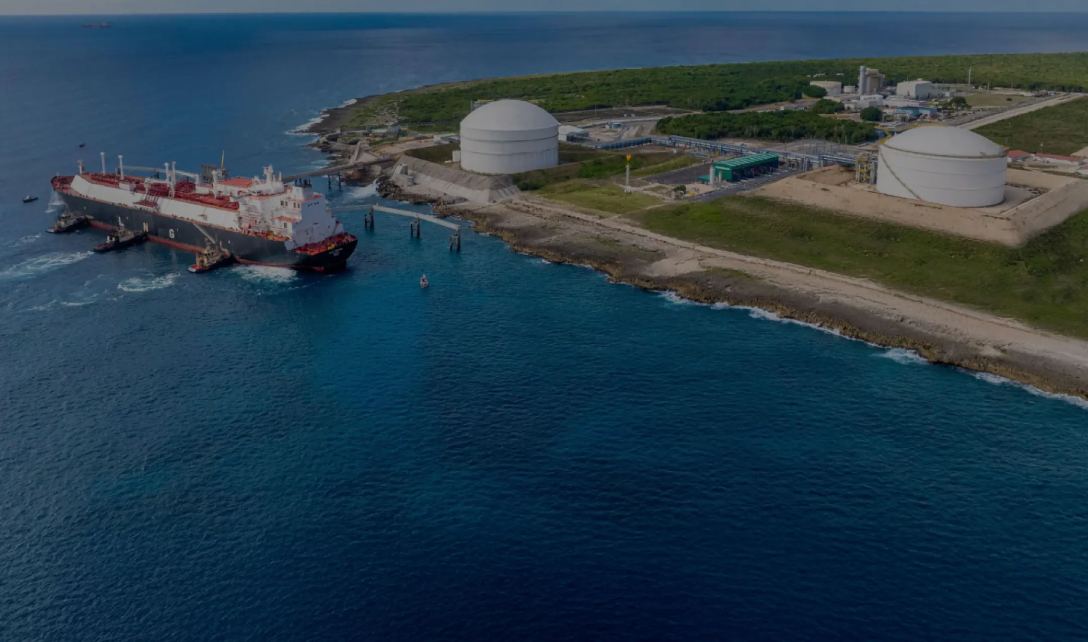 Aerial view of a large LNG tanker docked at a coastal terminal with two large storage tanks on a grassy area, surrounded by blue ocean water.