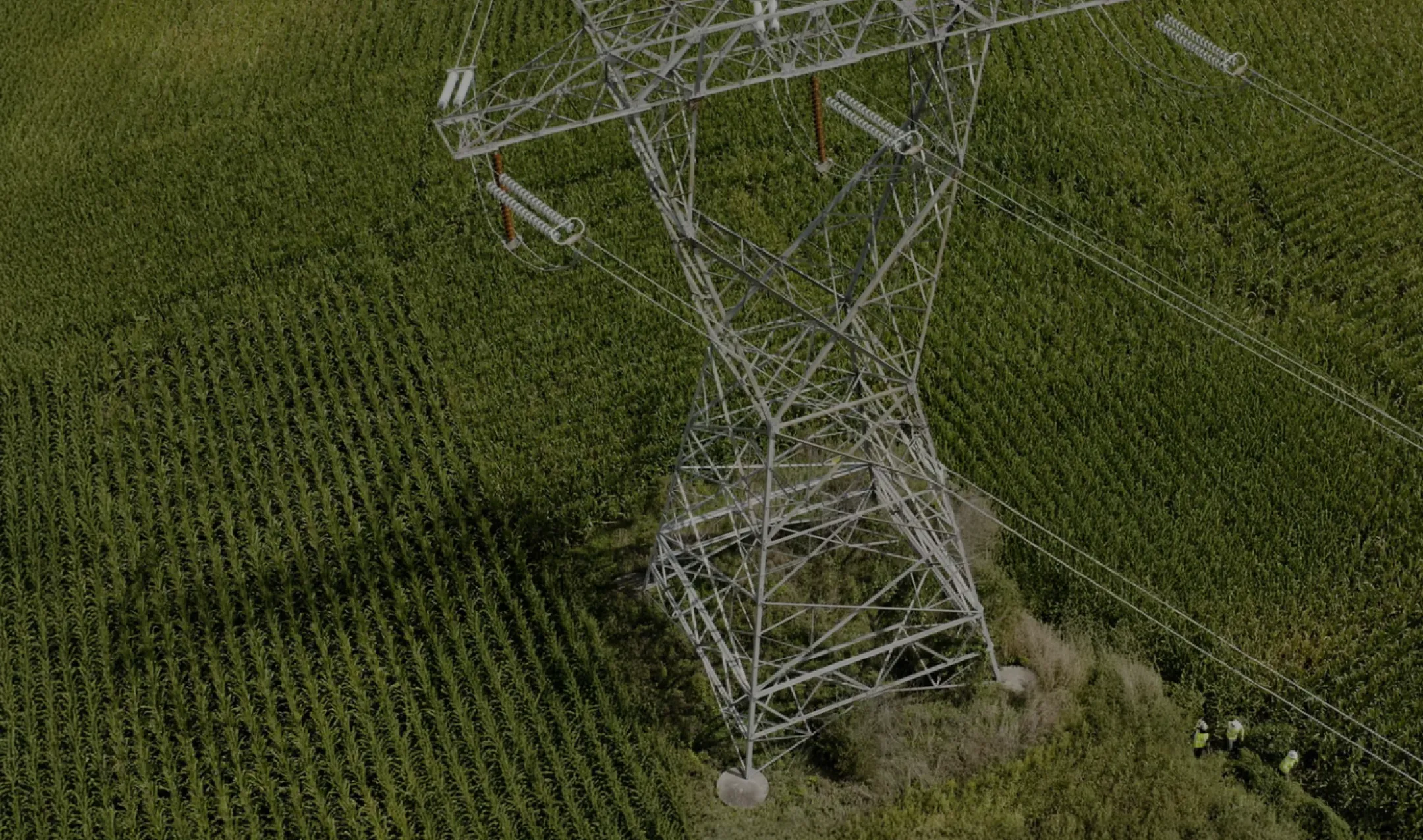 Aerial view of a large metal electricity pylon standing in the middle of a green agricultural field. Three workers in safety vests are visible near the base of the pylon.