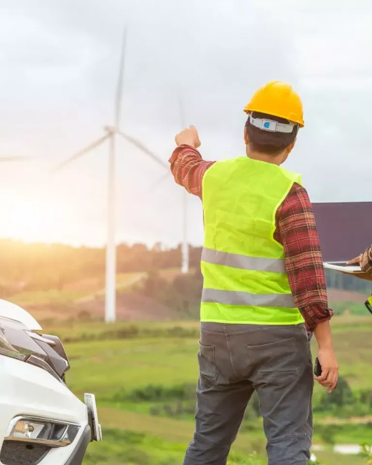 A person wearing a yellow hard hat and reflective vest points towards wind turbines in a grassy landscape. The sun is setting in the background, casting a warm glow over the scene. A white vehicle is partially visible on the left side.
