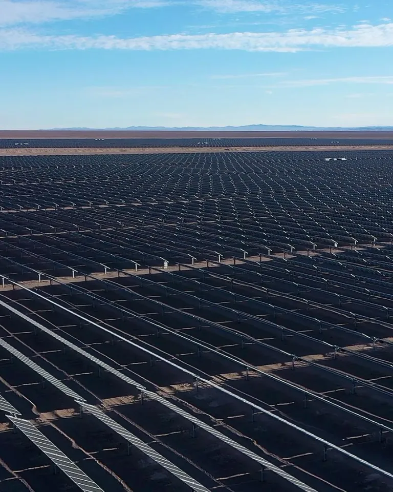 A vast solar farm with rows of solar panels stretching into the distance under a clear blue sky. The panels are arranged in parallel lines across a flat, barren landscape, with mountains visible on the horizon.