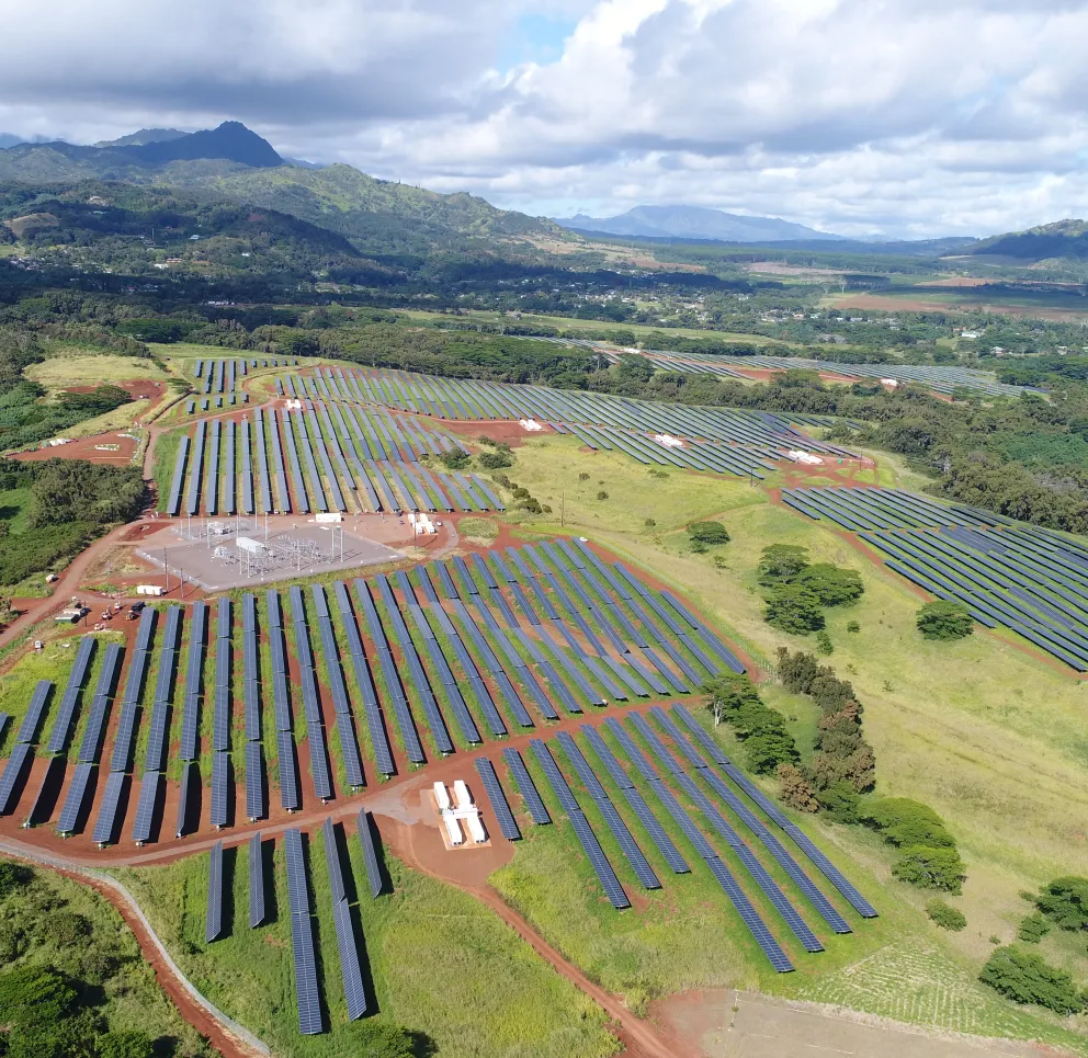 Aerial view of a large solar farm with rows of solar panels set on a hillside, surrounded by lush green vegetation and mountains in the background under a partly cloudy sky.