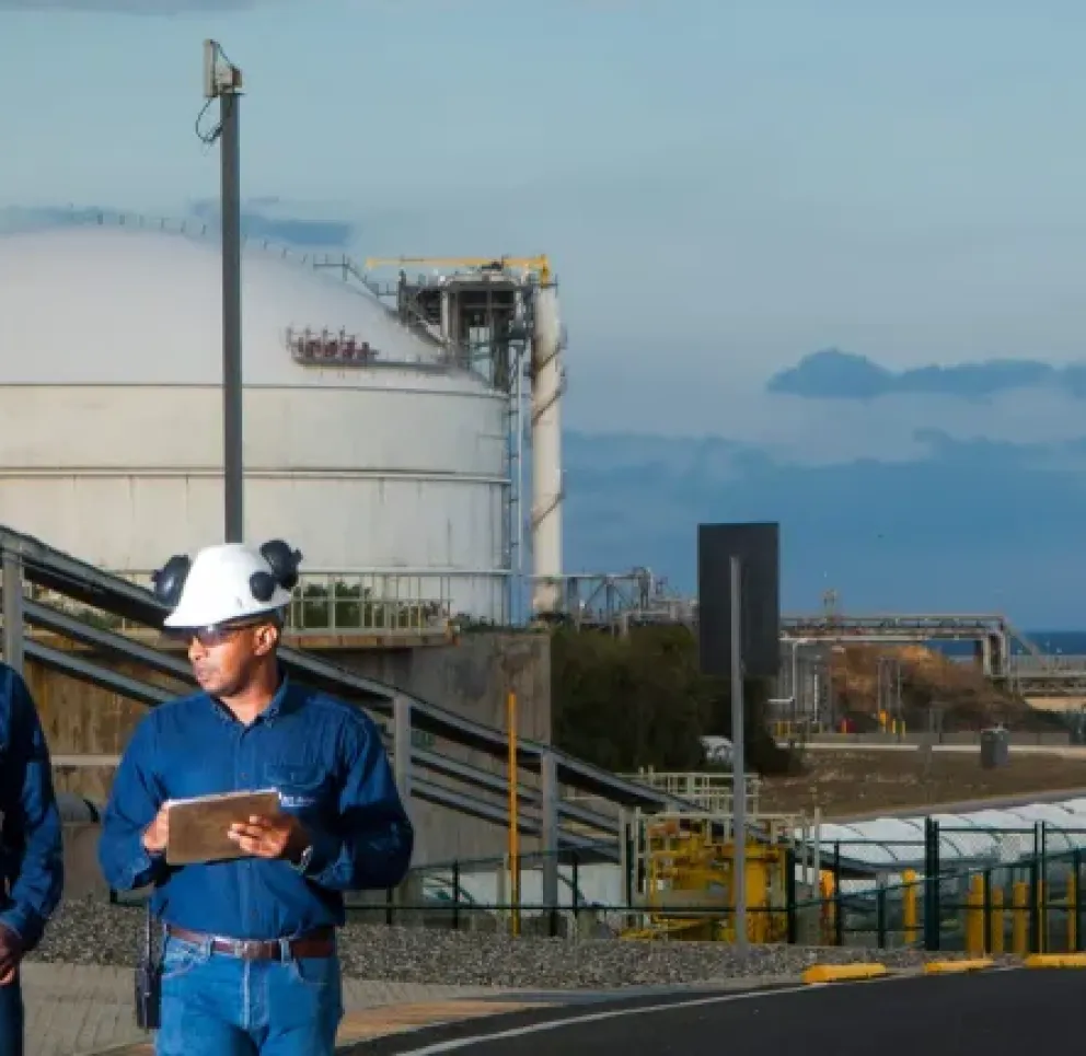 Two workers wearing blue shirts and white hard hats walk along a paved road at an industrial site. A large white storage tank is visible in the background, along with other industrial structures and pipes. The sky is partly cloudy.