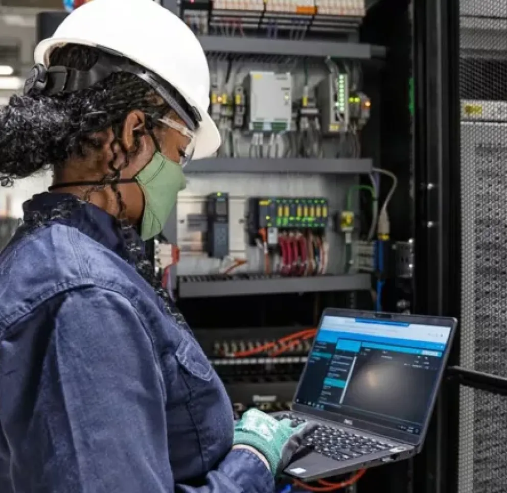 A person wearing a hard hat and face mask works on a laptop in a server room. They are standing in front of open server racks with visible wiring and equipment.