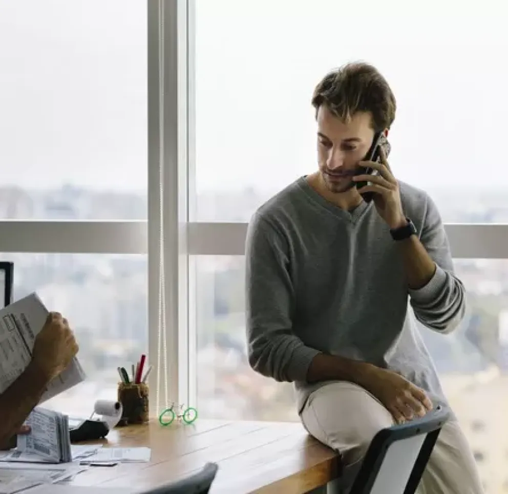 Two men in an office with large windows. One is sitting at a desk reviewing documents, while the other is leaning against the window, talking on a phone.
