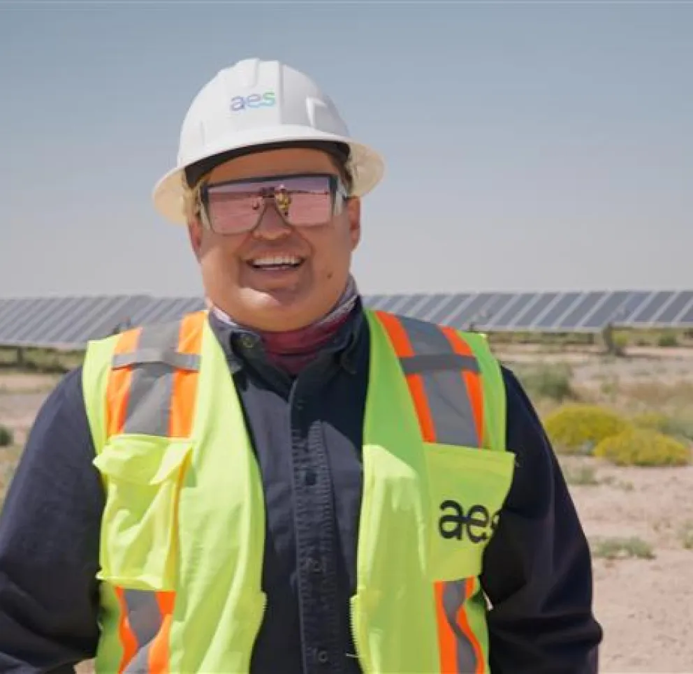 A person wearing a hard hat, safety glasses, and a high-visibility vest stands smiling in front of a field of solar panels on a sunny day.