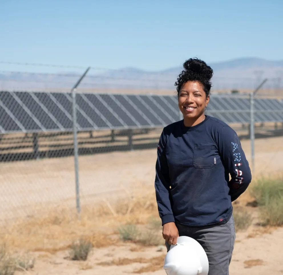 A person in a dark shirt holding a white hard hat stands in front of a solar panel array, with a fence and mountains in the background.