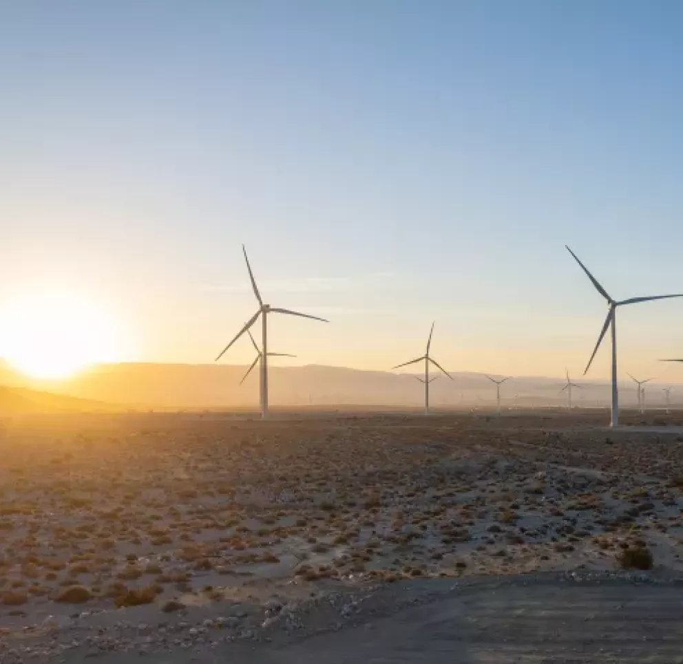 A landscape of a wind farm at sunrise, featuring multiple wind turbines scattered across a desert area. The sun is rising over distant hills, casting a warm glow across the sky and land.